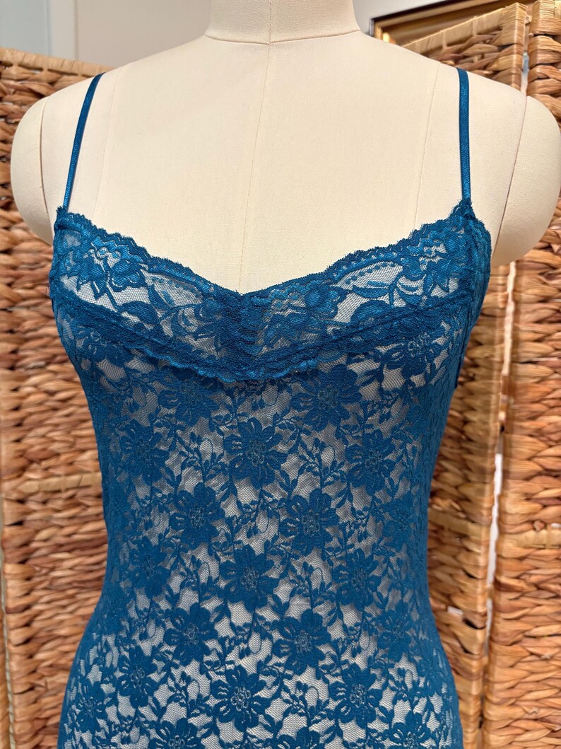 K&ouml;nnte beinhalten: Ein tealfarbenes Spitzen-Camisole mit zarten Spaghettitr&auml;gern. Das Camisole hat ein florales Spitzenmuster und einen gewellten Ausschnitt. Das Kleidungsst&uuml;ck wird auf einer Schaufensterpuppe pr&auml;sentiert.