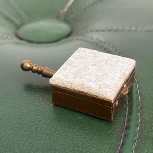 May include: A small, rectangular antique box with a brass handle and trim. The lid is covered in a mosaic of small, white, iridescent pieces. The box is resting on a green, tufted surface.