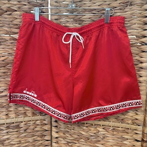 May include: Bright red athletic shorts with a white drawstring and a decorative geometric pattern along the hem. The shorts are hanging on a metal hanger against a woven background.