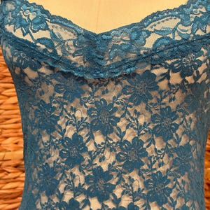 K&ouml;nnte beinhalten: Ein tealfarbenes Spitzen-Camisole mit floralem Muster. Das Camisole hat zarte Spaghettitr&auml;ger und eine gewellte Spitzenborte am Ausschnitt. Das filigrane florale Design bedeckt das gesamte Kleidungsst&uuml;ck und erzeugt ein strukturiertes Aussehen.