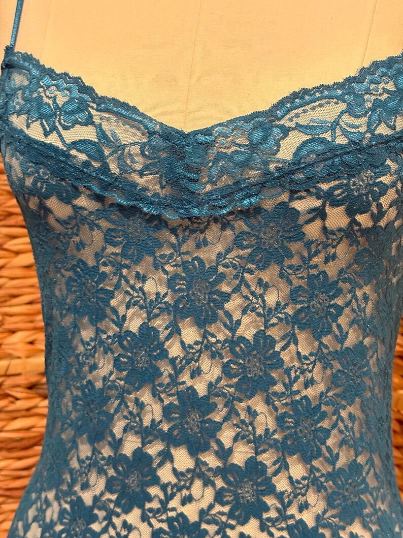 K&ouml;nnte beinhalten: Ein tealfarbenes Spitzen-Camisole mit floralem Muster. Das Camisole hat zarte Spaghettitr&auml;ger und eine gewellte Spitzenborte am Ausschnitt. Das filigrane florale Design bedeckt das gesamte Kleidungsst&uuml;ck und erzeugt ein strukturiertes Aussehen.