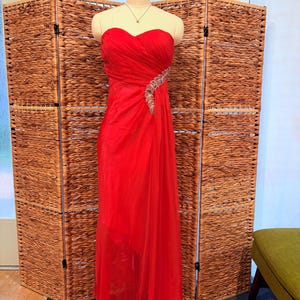 May include: A strapless, floor-length red gown on a mannequin. The bodice is ruched, and a beaded embellishment accents the waist. The skirt drapes elegantly, and the dress is displayed in front of a woven room divider.