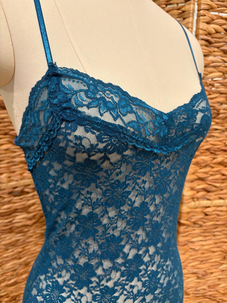 K&ouml;nnte beinhalten: Ein teal-farbener Body aus floraler Spitze mit d&uuml;nnen Spaghettitr&auml;gern. Die zarte Spitze hat einen gewellten Rand am Ausschnitt und ein Blumenmuster auf dem gesamten Kleidungsst&uuml;ck. Der Body wird auf einer Schaufensterpuppe pr&auml;sentiert.