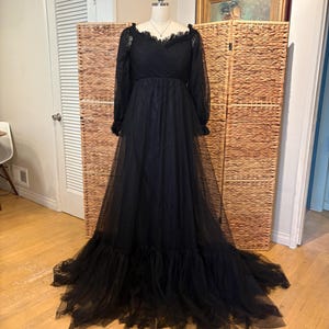 May include: A long, black formal gown with a tulle skirt and long sleeves. The dress features a ruffled neckline and cuffs, and a flowing train. The dress is displayed on a mannequin in front of a woven screen.