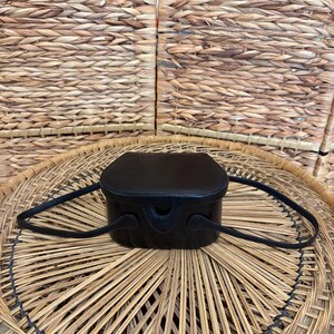 May include: A small, black leather handbag with a rounded rectangular shape and a top handle. The bag has a structured design and is set against a woven background. The bag's dimensions are approximately 6 inches in width and 5 inches in height.