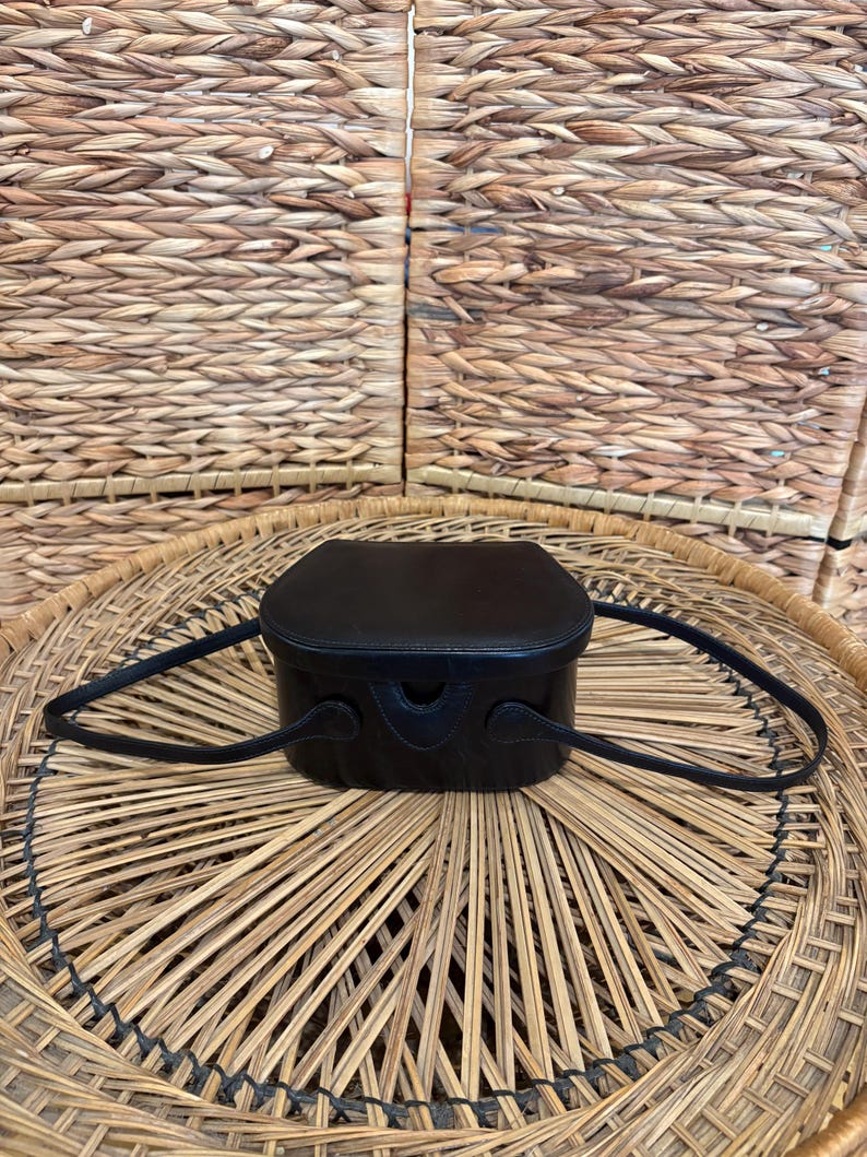 May include: A small, black leather handbag with a rounded rectangular shape and a top handle. The bag has a structured design and is set against a woven background. The bag's dimensions are approximately 6 inches in width and 5 inches in height.