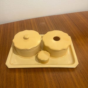 May include: A vintage vanity set in a pale yellow color. The set includes two round lidded containers, a small round container with a lid, and a rectangular tray. The containers have a scalloped edge design.