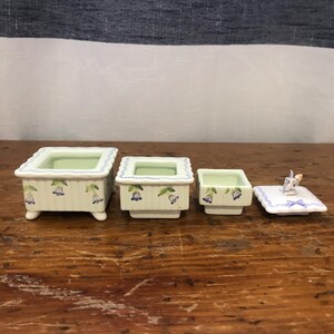 May include: A set of four porcelain trinket boxes. The boxes are rectangular with a light green interior and a cream exterior decorated with blue floral designs. The smallest box has a lid with a butterfly figurine.