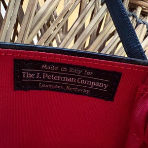 May include: Close-up of a black label inside a red handbag. The label reads "Made in Italy for The J. Peterman Company, Lexington, Kentucky." The bag has a black strap and a red interior.