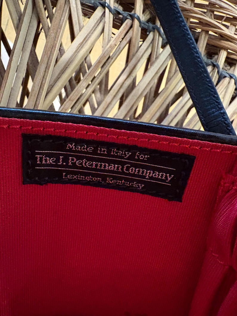 May include: Close-up of a black label inside a red handbag. The label reads "Made in Italy for The J. Peterman Company, Lexington, Kentucky." The bag has a black strap and a red interior.