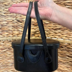 May include: Black leather handbag with two top handles. The rectangular bag has a curved front detail and a structured design. The handles are thin and attached to the top of the bag. The background is a woven texture.
