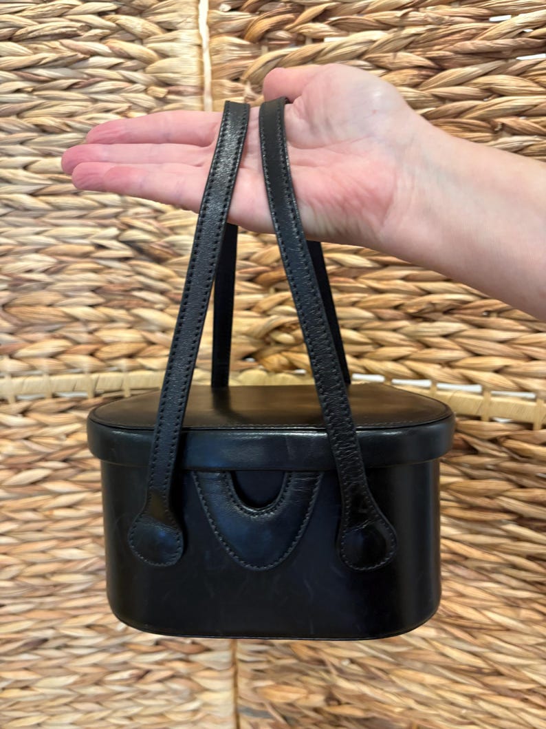 May include: Black leather handbag with two top handles. The rectangular bag has a curved front detail and a structured design. The handles are thin and attached to the top of the bag. The background is a woven texture.