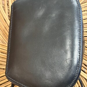 May include: Black leather crossbody bag with a rounded top and a stitched border. The bag has a long, thin strap and appears to be made of smooth leather. The bag is set against a light brown woven background.