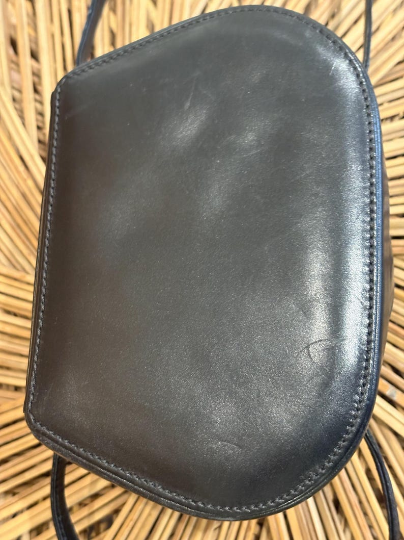 May include: Black leather crossbody bag with a rounded top and a stitched border. The bag has a long, thin strap and appears to be made of smooth leather. The bag is set against a light brown woven background.