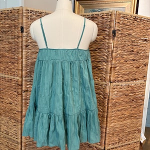 Op de afbeelding: Een teal-kleurige, gelaagde jurk met dunne spaghettibandjes. De jurk wordt getoond op een mannequin, waarbij de gerimpelde lagen en de textuur van de stof worden getoond. De jurk heeft een relaxte, vloeiende silhouet.