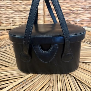 May include: Black leather handbag with a rectangular body and a rounded top. The bag features two short handles and a small, curved opening on the front. The bag is sitting on a woven surface.
