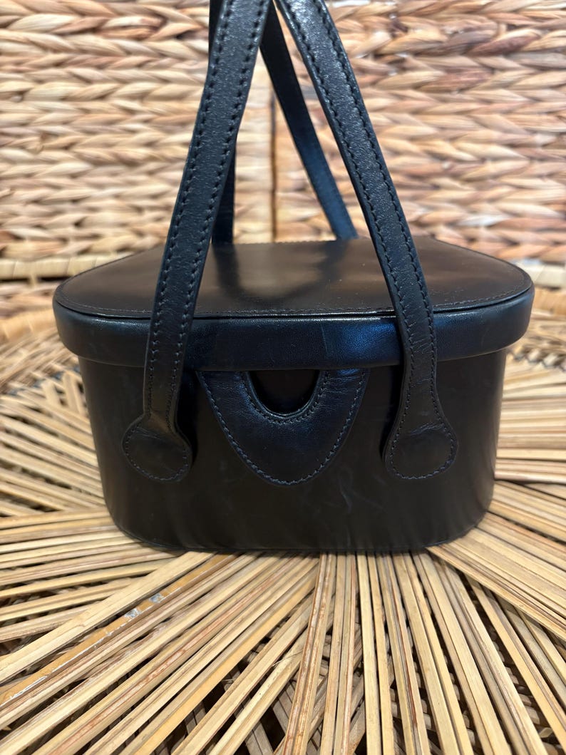 May include: Black leather handbag with a rectangular body and a rounded top. The bag features two short handles and a small, curved opening on the front. The bag is sitting on a woven surface.