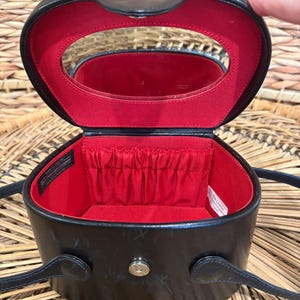May include: Black oval-shaped cosmetic case with a red interior. The case has a mirror inside the lid and a small pocket. It features a handle and a shoulder strap. The case is open, revealing the interior.