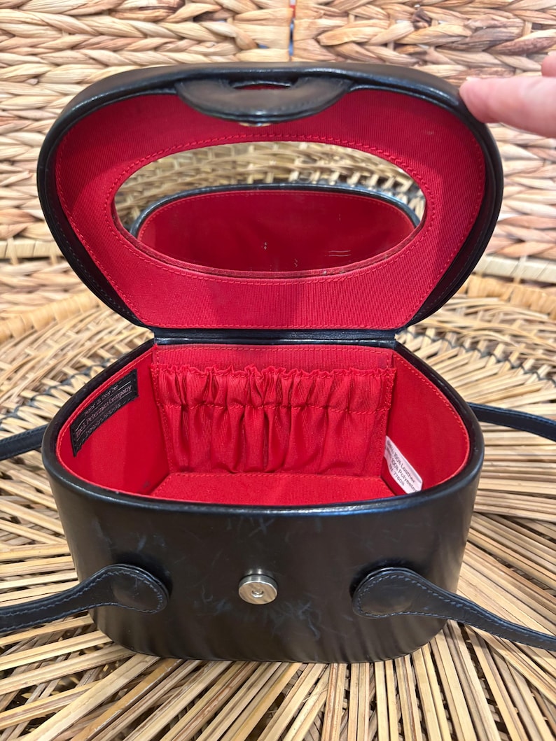 May include: Black oval-shaped cosmetic case with a red interior. The case has a mirror inside the lid and a small pocket. It features a handle and a shoulder strap. The case is open, revealing the interior.