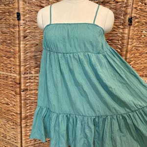 Op de afbeelding: Een turquoisekleurige, gelaagde jurk met dunne spaghettibandjes. De jurk heeft een vierkante halslijn en een gefranjerde zoom. De stof lijkt licht en getextureerd, met verticale lijnen. De jurk wordt op een mannequin getoond.