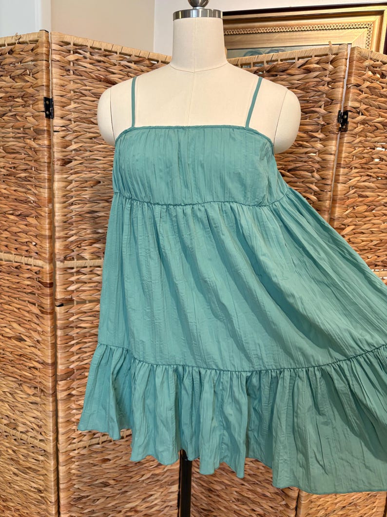 Op de afbeelding: Een turquoisekleurige, gelaagde jurk met dunne spaghettibandjes. De jurk heeft een vierkante halslijn en een gefranjerde zoom. De stof lijkt licht en getextureerd, met verticale lijnen. De jurk wordt op een mannequin getoond.