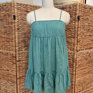 Op de afbeelding: Een turquoise jurk met dunne spaghettibandjes en een gelaagde, gerimpelde rok. De jurk wordt gepresenteerd op een mannequin tegen een geweven achtergrond. De jurk heeft een getextureerd, geplooid lijfje.