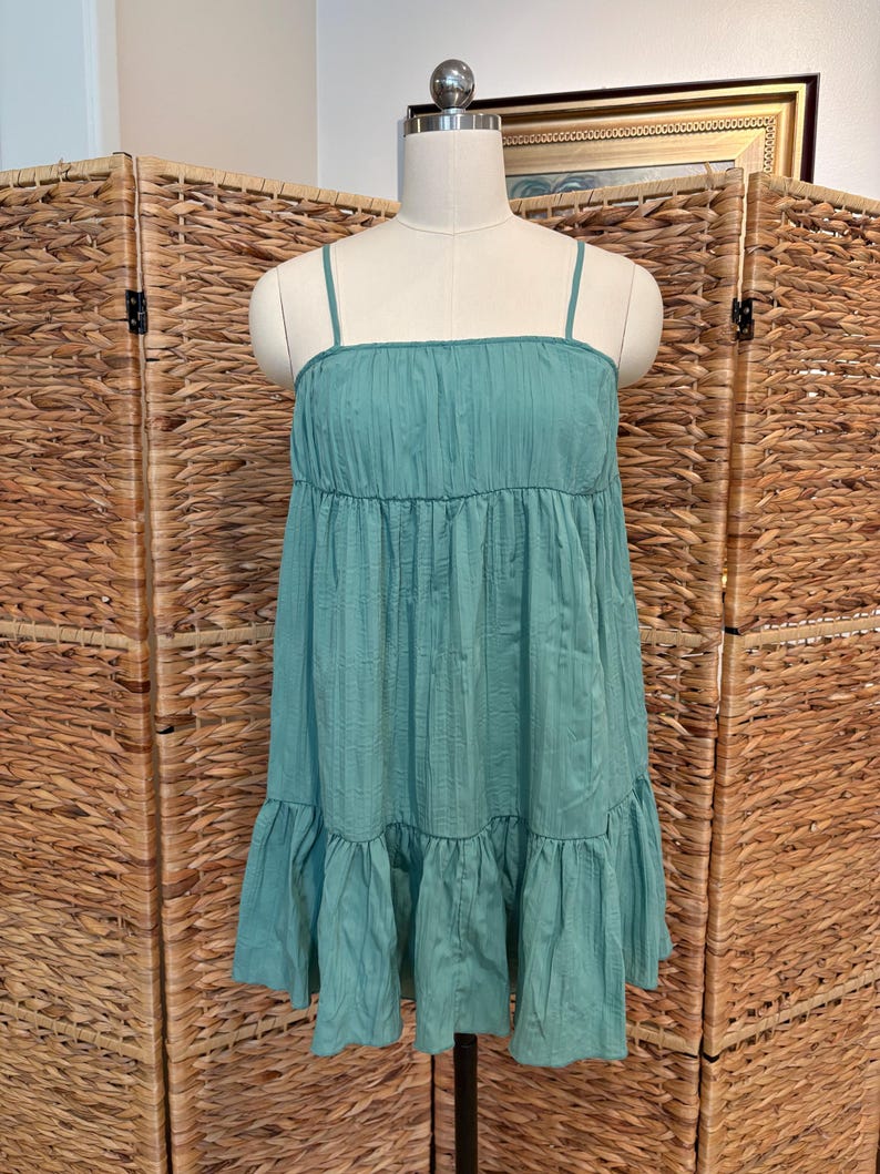Op de afbeelding: Een turquoise jurk met dunne spaghettibandjes en een gelaagde, gerimpelde rok. De jurk wordt gepresenteerd op een mannequin tegen een geweven achtergrond. De jurk heeft een getextureerd, geplooid lijfje.