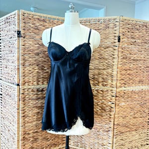 Könnte beinhalten: Schwarzes Satin-Slip mit herzförmigem Ausschnitt und zarter Spitzenborte. Die Dessous werden auf einer Schaufensterpuppe vor neutralem Hintergrund präsentiert. Das Slip hat verstellbare Träger und ein figurbetontes Oberteil.