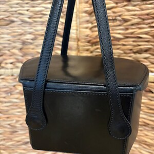 May include: A black leather handbag with a rectangular shape and a top handle. The bag has a structured design with visible stitching and a smooth, glossy finish. The handle is attached with a rounded, decorative piece. The background is a woven texture.