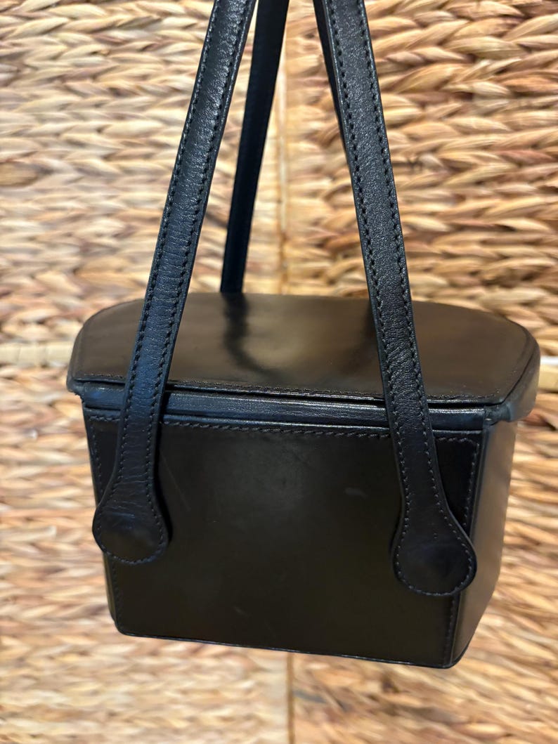 May include: A black leather handbag with a rectangular shape and a top handle. The bag has a structured design with visible stitching and a smooth, glossy finish. The handle is attached with a rounded, decorative piece. The background is a woven texture.