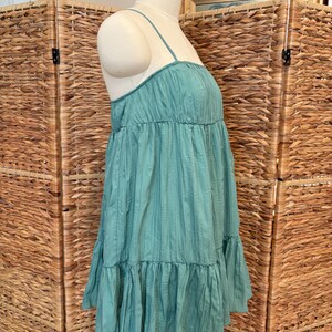 Op de afbeelding: Een turquoise jurk met dunne spaghettibandjes en een gelaagde, gerimpelde rok. De jurk heeft een textuur, verticaal gestreept patroon en wordt op een mannequin getoond. De jurk is een casual zomerstijl.
