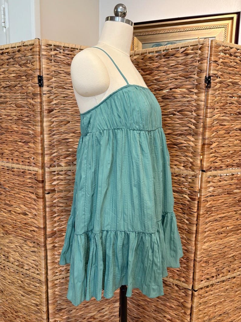 Op de afbeelding: Een turquoise jurk met dunne spaghettibandjes en een gelaagde, gerimpelde rok. De jurk heeft een textuur, verticaal gestreept patroon en wordt op een mannequin getoond. De jurk is een casual zomerstijl.