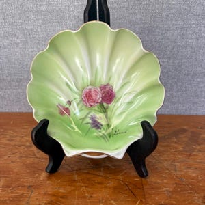 May include: A scalloped, light green ceramic dish with a floral design featuring pink and purple flowers. The dish has a gold rim and is displayed on a black stand. The background is a light gray.