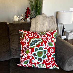 May include: Red, white, and teal floral patterned throw pillow with a brown couch in the background.