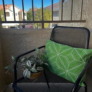 May include: A black metal chair with a green and white geometric patterned pillow. A potted plant with green and white leaves sits on the chair's armrest. The chair is on a balcony overlooking a view of buildings and trees.