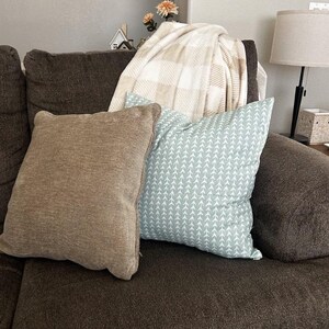 May include: A brown couch with three throw pillows. The pillows are brown, light blue with a white arrow pattern, and a white and beige plaid. A throw blanket is draped over the back of the couch.