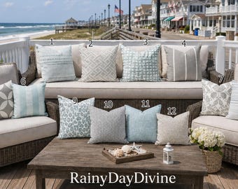 May include: A collection of twelve square throw pillows featuring various patterns including stripes, geometric shapes, and animal prints in shades of blue, gray, and white. The pillows are arranged on outdoor furniture on a deck.