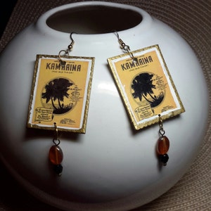 May include: A pair of handmade earrings featuring rectangular pendants with a vintage design. The pendants have a yellow background with black text and a palm tree silhouette. Each earring has a gold-colored border and a dangling bead.