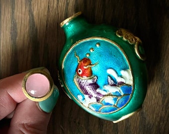 May include: A small, green, round, decorative bottle with a gold lid and a blue and white enamel design of a fish. The bottle is on a wooden surface.