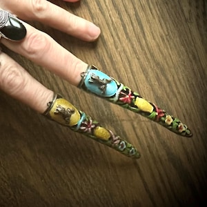 Peut inclure: Une main avec de longs ongles peints. Les ongles sont décorés de motifs colorés et de petites touches dorées. La main porte une bague en argent avec une pierre noire.