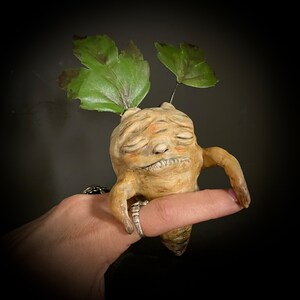 May include: A hand holds a small, whimsical sculpture of a humanoid figure with closed eyes and a peaceful expression. The figure is made of brown clay and has two leafy green appendages growing from its head.
