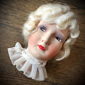 May include: A vintage doll head with painted facial features, including red lips, rosy cheeks, and detailed eye makeup. The doll has blonde, curly hair and wears a white lace collar. The head is resting on a wooden surface.