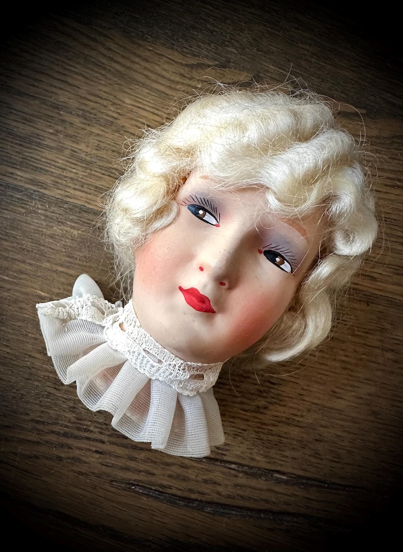 May include: A vintage doll head with painted facial features, including red lips, rosy cheeks, and detailed eye makeup. The doll has blonde, curly hair and wears a white lace collar. The head is resting on a wooden surface.