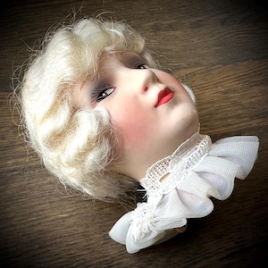 May include: A vintage doll head with blonde, wavy hair, rosy cheeks, and red lips. The doll head has detailed facial features and is wearing a white lace collar. The doll head is lying on a wooden surface.