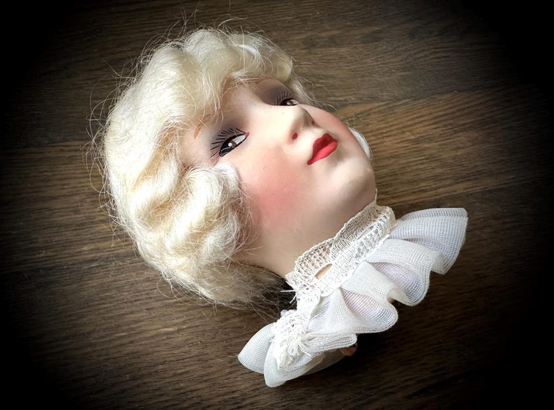 May include: A vintage doll head with blonde, wavy hair, rosy cheeks, and red lips. The doll head has detailed facial features and is wearing a white lace collar. The doll head is lying on a wooden surface.