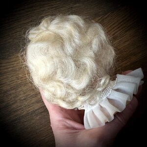 May include: A doll wig with light blonde, wavy hair. The wig is attached to a white, ruffled fabric collar. The hair is styled in loose curls. The background is a dark wooden surface.
