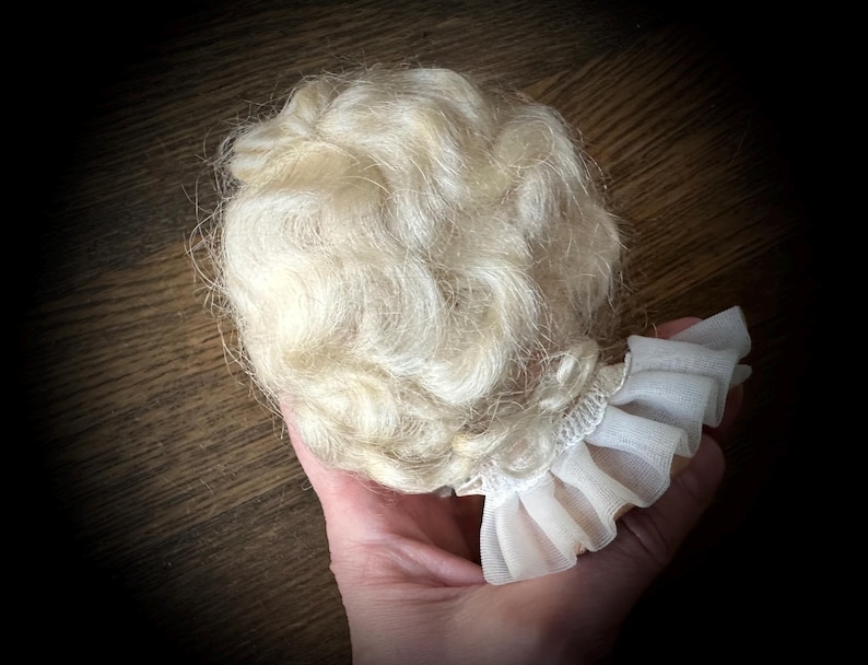 May include: A doll wig with light blonde, wavy hair. The wig is attached to a white, ruffled fabric collar. The hair is styled in loose curls. The background is a dark wooden surface.