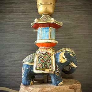 Könnte beinhalten: Dekorative Lampe mit blauem Elefantenfuß und goldenen Details, gekrönt von einer Pagodenstruktur. Die Lampe hat eine Messingfassung und steht auf einem Holzsockel. Der Elefant ist mit floralen Mustern verziert.