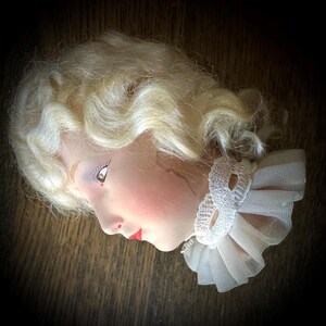 May include: A vintage doll head with blonde hair styled in curls. The doll has a painted face with red lips and a white lace and tulle collar. The head is set against a dark background.