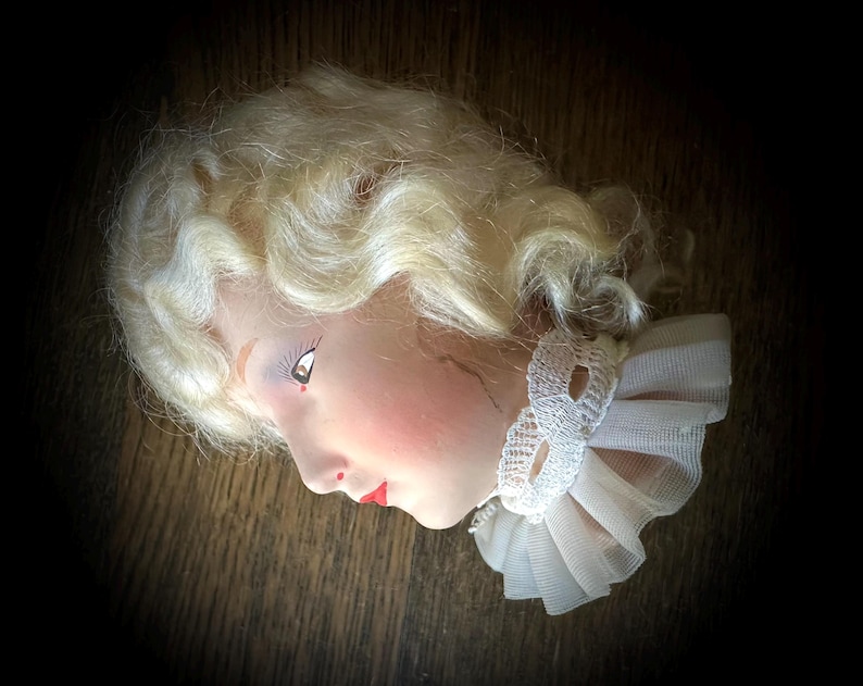 May include: A vintage doll head with blonde hair styled in curls. The doll has a painted face with red lips and a white lace and tulle collar. The head is set against a dark background.